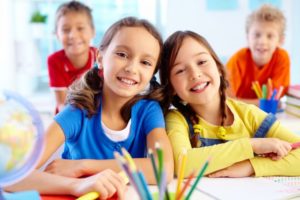 2 boys blurred in the background, two girls in foreground, smiling, sitting in class at their desk